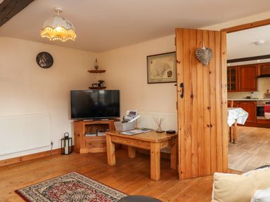 A living room with a television on a wooden stand a wooden coffee table and an open wooden door showing a kitchen at Northwood Barn in Okehampton