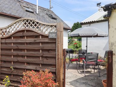 A small outdoor patio with a table and four chairs under a grey umbrella behind a wooden gate at The Barn in Okehampton