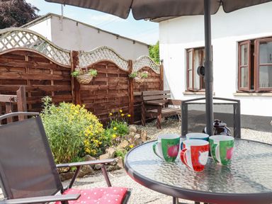 A garden patio with a glass table holding colorful mugs and a black umbrella over the table at Northwood Barn in Okehampton