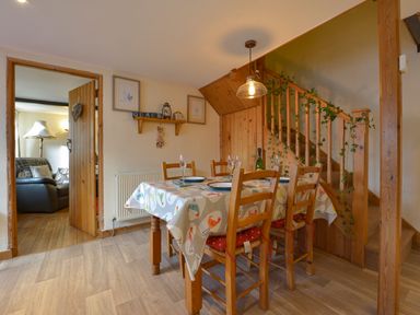 A dining area with a table set for four, wooden chairs, a staircase with ivy decoration, and an open door leading to a living room at Northwood Barn in Okehampton
