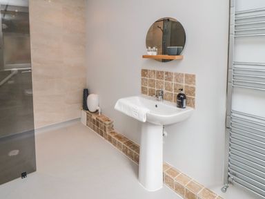 A bathroom with a pedestal sink towel soap dispenser round mirror tiled wall and glass shower at The Annexe in Bridlington