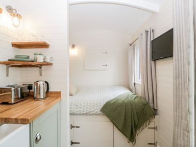 A small room with a bed, a wall-mounted TV, curtains, wooden shelves with dishes, a toaster, and a kettle at Willow in Kemble