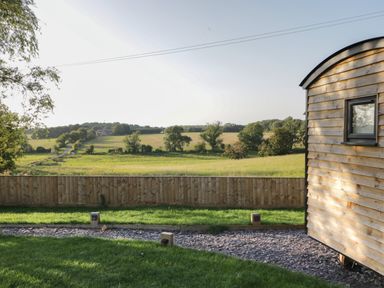 A wooden fence and grass lawn with a wooden structure on the right and fields with trees in the background at Willow in Kemble