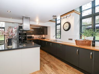 A kitchen with dark cabinets wooden countertops a clock on the wall and windows overlooking a garden at Owl Barn in Marston Stannett near Leominster