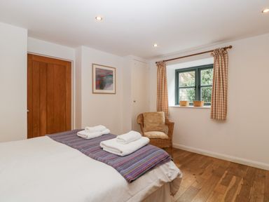 A bedroom with a bed topped with folded towels a wicker chair by a window with checkered curtains and wooden door at Owl Barn Marston Stannett near Leominster