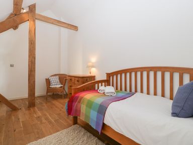 A bedroom with a wooden bed a rainbow blanket a wooden chest of drawers and a wicker chair at Owl Barn in Marston Stannett near Leominster