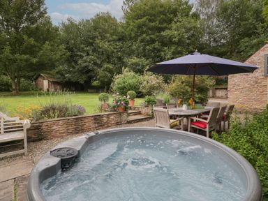 An outdoor patio with a bubbling hot tub wooden table and chairs under an umbrella and a garden with trees and plants at Owl Barn Marston Stannett near Leominster