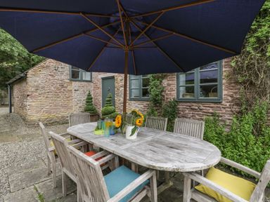 An outdoor dining table with six wooden chairs and a large umbrella on a stone patio at Owl Barn Marston Stannett near Leominster