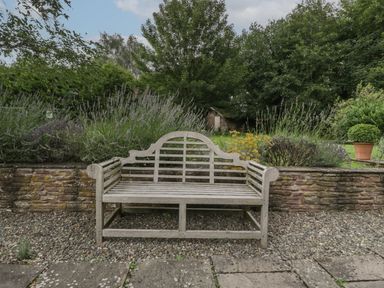 A wooden bench on gravel in front of a stone wall with plants and a small wooden shed behind at Owl Barn Marston Stannett near Leominster