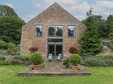 The rear exterior of a stone house with two wooden chairs on a patio surrounded by potted plants and shrubbery at Owl Barn in Marston Stannett near Leominster