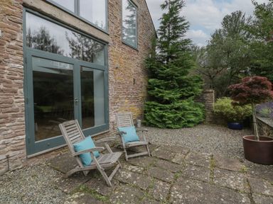 A stone patio with two wooden chairs and blue cushions outside a building with large windows at Owl Barn in Marston Stannett near Leominster
