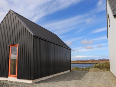 A black house with a red window frame beside a white building near a body of water at North Light Studio in Locheport near Lochmaddy North Uist