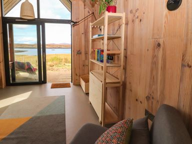 A room with wooden walls a shelf with books a gray chair and a sliding glass door overlooking a waterfront at North Light Studio in Locheport near Lochmaddy North Uist