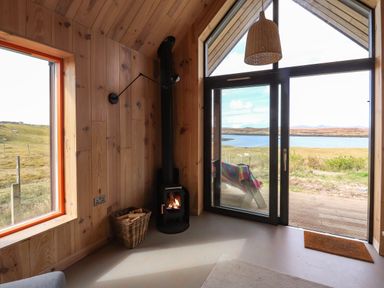 A room with wooden walls featuring a wood-burning stove a basket with firewood large windows and a glass door overlooking a lake at North Light Studio Locheport near Lochmaddy North Uist
