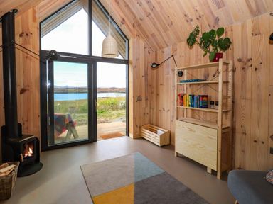 A room with wooden walls large glass doors a burning wood stove bookshelf and a window overlooking a body of water at North Light Studio in Locheport near Lochmaddy North Uist