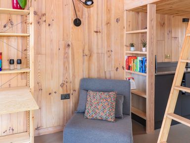 A small room with wooden walls featuring a gray chair with two pillows a bookshelf with colorful books and plants a fold-down wooden desk and a ladder by the kitchen area at North Light Studio in Locheport near Lochmaddy North Uist