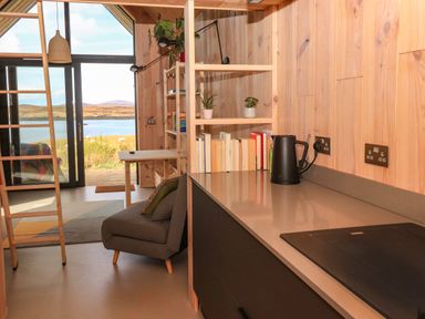 A small living area with a gray chair and bookshelves next to a kitchen countertop and sliding glass doors overlooking a water body at North Light Studio in Locheport near Lochmaddy North Uist