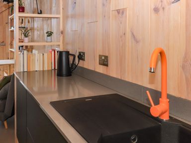 A kitchen countertop with an orange faucet a black sink a black electric kettle books on a shelf and wooden wall panels at North Light Studio in Locheport near Lochmaddy North Uist
