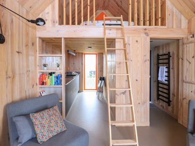 A wooden interior with a small kitchen, a ladder leading to a loft bed, grey chairs with cushions, and a bookshelf with books at North Light Studio Locheport near Lochmaddy North Uist
