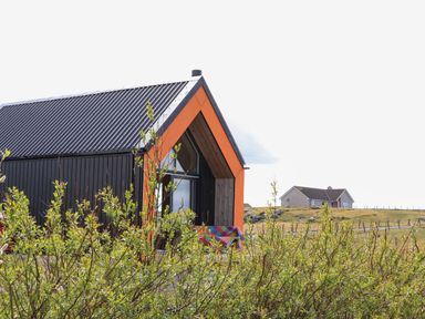 Modern house with black walls and orange framing near green bushes and open field at North Light Studio in Locheport near Lochmaddy North Uist
