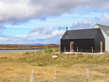 A black house with a sloped roof next to a body of water with grassy land and sheep around at North Light Studio in Locheport near Lochmaddy North Uist