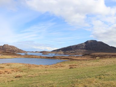 A landscape with grassy fields lakes and rocky hills under a partly cloudy sky at North Light Studio in Locheport near Lochmaddy North Uist
