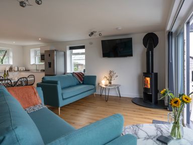 A living room with blue sofas a wood burning stove a wall mounted tv and an open kitchen at Lanyon in St Ives