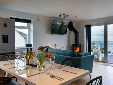 A dining table set with wine bottles and glasses next to a living room with a green sofa and a wood stove at Lanyon in St Ives