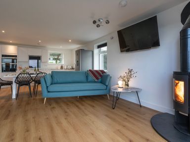An open living area with a blue sofa next to a small table with flowers and a lamp a wall-mounted TV a fireplace and a kitchen with a dining table and chairs at Lanyon in St Ives