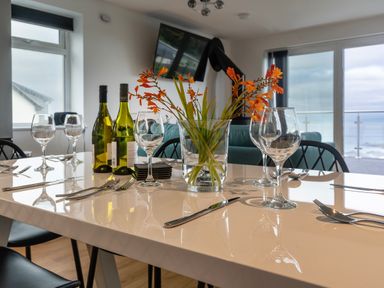 A dining table set with wine glasses cutlery and a vase of orange flowers in a room with large windows and a television at Lanyon in St Ives