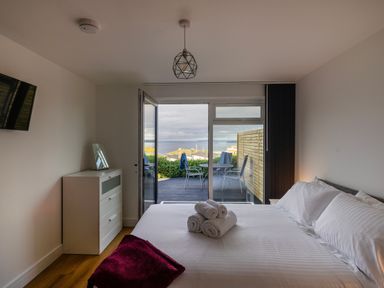 A bedroom with a bed towels and a dresser opening onto a patio with chairs and a sea view at Lanyon in St Ives