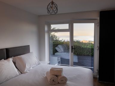 A bedroom with a bed with white linens and rolled towels in front of glass doors overlooking greenery at Lanyon in St Ives
