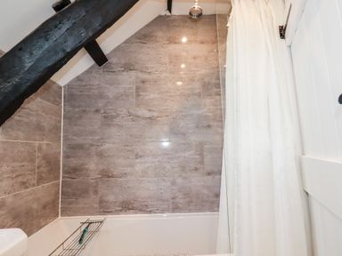 A bathroom with a bathtub shower combination stone tiled walls a white shower curtain and a black wooden ceiling beam at Ivy Cottage in Trenale near Tintagel