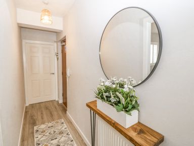 A hallway with a wooden console table holding a white planter with green plants and white flowers and a round mirror on the wall above at Sea La Vie in Benllech