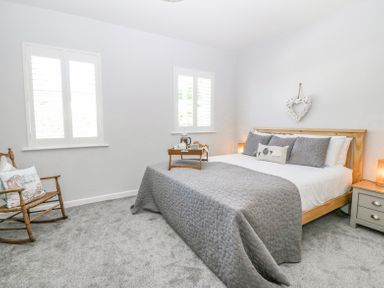 A bedroom with a wooden bed and gray bedding by two windows and a rocking chair at Sea La Vie in Benllech
