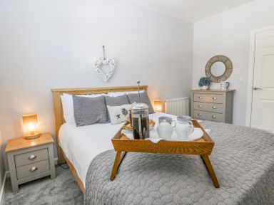A bedroom with a wooden bed frame with gray pillows and bedding a wooden tray with a coffee press and cups a gray nightstand with a lamp and a gray chest of drawers with a round mirror at Sea La Vie in Benllech