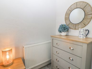 A corner with a chest of drawers a round mirror a lamp and a vase with flowers at Sea La Vie in Benllech