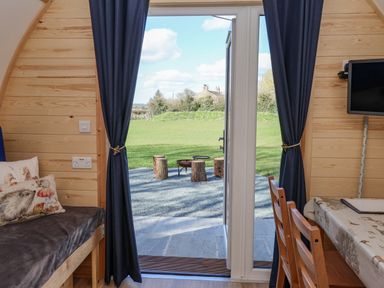 A wooden interior room with a sofa and pillows on the left two wooden chairs and a table with cloth on the right and an open door leading to an outdoor area with a fire pit and log stools at Mischief in Little Kelk near Bridlington