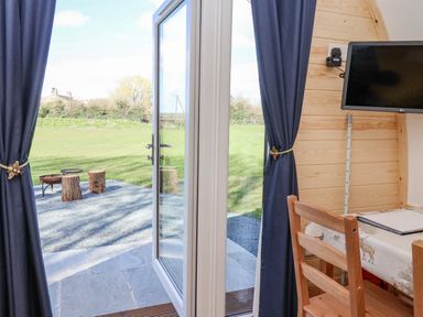 A wooden chair and a table with a decorative cloth next to a wall mounted tv and open glass door leading outside showing tree stump seats and grass at Mischief Little Kelk near Bridlington