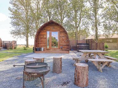 A wooden cabin with a glass door surrounded by trees with a fire pit and log stools in front and a wooden picnic table to the right at Mischief Little Kelk near Bridlington