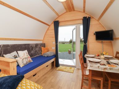 An interior view of a wooden pod with a blue cushioned bench, dining table with chairs, open door leading outside and a wall-mounted TV at Covey in Little Kelk near Bridlington