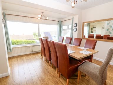 A dining room with a wooden table surrounded by red and beige chairs next to large windows at Willoway in Benllech