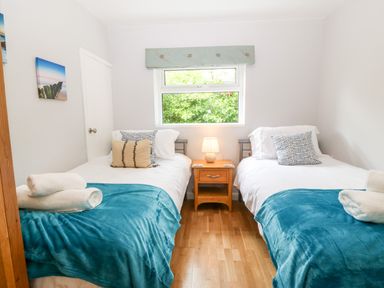 A bedroom with two single beds with blue blankets and pillows a wooden nightstand with a lamp and a window showing greenery at Willoway in Benllech