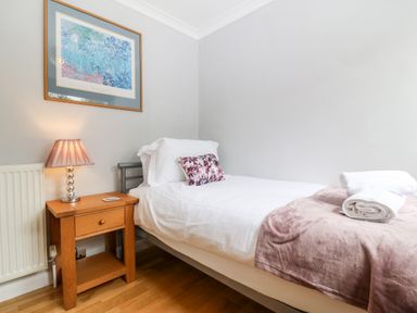 A single bed with white linens and a purple pillow next to a wooden bedside table with a lamp and a framed picture on a wall at Willoway in Benllech