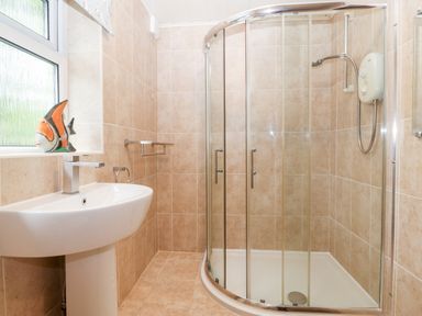 A bathroom with a curved glass shower cubicle a white sink with a modern faucet and a window with a fish decoration on the sill at Willoway in Benllech