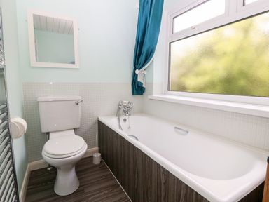 A bathroom with a toilet a bathtub with patterned sides a small mirror and a window with a blue curtain at Willoway in Benllech