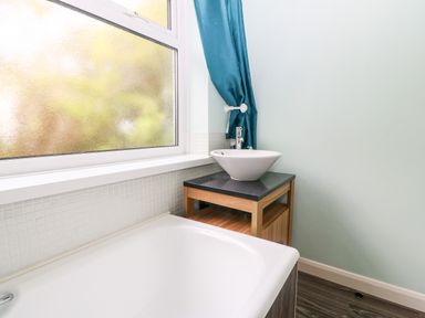 A bathroom with a bathtub a basin on a wooden cabinet and a frosted window with a blue curtain at Willoway in Benllech