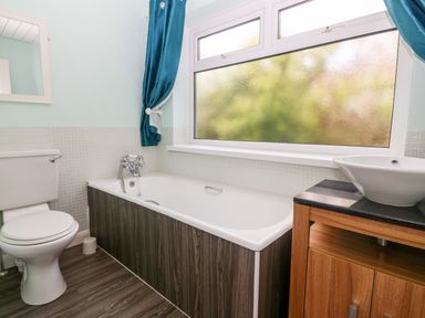 A bathroom with a toilet a bathtub with wooden paneling a window with blue curtains and a wooden sink cabinet at Willoway Benllech