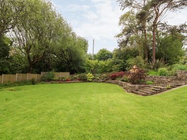 A lawn area with wooden steps and a stone retaining wall surrounded by trees and shrubs at Willoway in Benllech