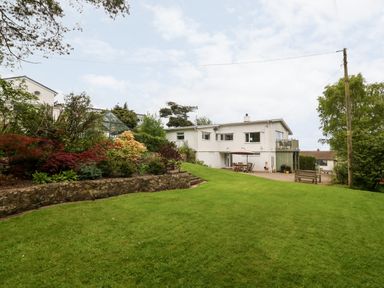 A backyard with green lawn and raised flower bed and a white house with patio furniture and umbrella at Willoway in Benllech